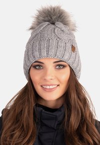 Berretto grigio a maglia con un grande pompon in pelliccia artificiale sulla parte superiore, caratterizzato da un motivo a treccia testurizzato e una piccola etichetta in pelle.