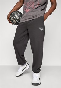 Person med mørke joggers med et sølvfalk-logo, hvide sneakers, holder en sort Spalding basketball og en grafisk t-shirt.