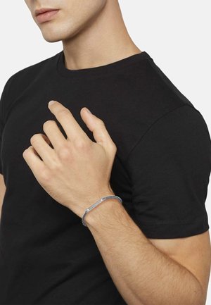 Silbernes Armband mit einem strukturierten Kettendesign und einem eleganten, rechteckigen Verschluss; getragen an einer Hand vor einem schwarzen Hemd Hintergrund.