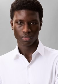 Hvid skjorte med klassisk krave og knaplukning foran, med blødt stof og en skræddersyet pasform, vist mod en grå baggrund.