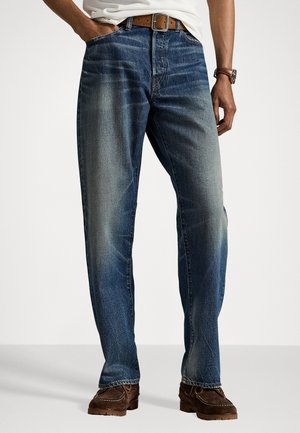 Uomo che indossa jeans blu sbiaditi con cintura marrone, camicia bianca, orologio marrone e scarpe scamosciate marroni scure, in piedi contro uno sfondo bianco.