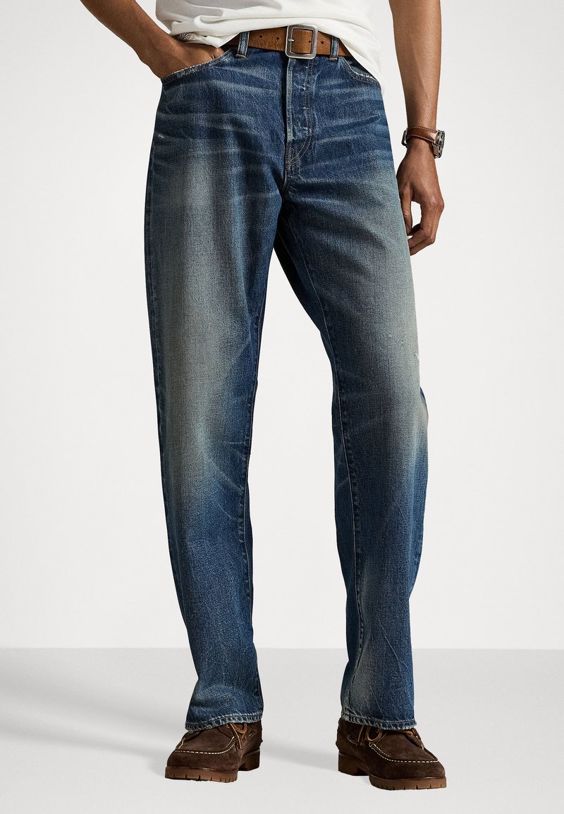 Man wearing faded blue jeans with brown belt, white shirt, brown watch, and dark brown suede shoes standing against white background.