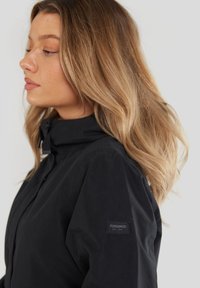Chaqueta negra con alto cuello y cierre de botón. Presenta un parche de marca en la manga izquierda. Textura suave y ajuste entallado.