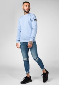 Sudadera azul claro con puños acanalados, combinada con jeans azul desgastados y zapatillas negras. Detalle notable del logo en la manga.
