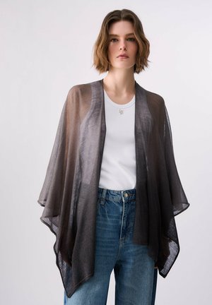 Mujer que lleva un chal negro translúcido sobre una camiseta sin mangas blanca con vaqueros azules, de pie frente a un fondo blanco liso.