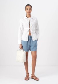 White button-up shirt with tie detail, denim shorts, light brown sandals, and a cream bag with a zigzag texture. Casual summer outfit.