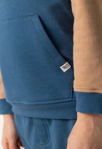 Felpa blu con maniche beige. Presenta una tasca frontale, polsini e orlo a costine, e un piccolo etichetta con logo sul lato. Materiale morbido in misto cotone.