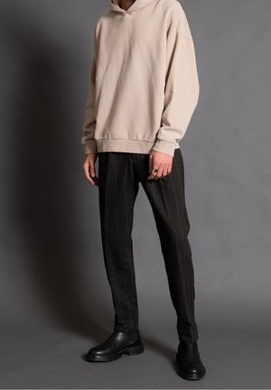 Person iført en beige hættetrøje, sorte bukser med nålestriber og sorte læderstøvler, stående mod en ensfarvet grå baggrund.