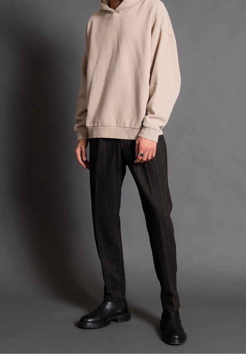 Personne portant un sweat à capuche beige, un pantalon noir à fines rayures et des bottes en cuir noires, debout devant un fond gris uni.