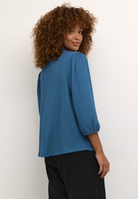 Haut bleu texturé avec manches bouffantes 3/4 et encolure ronde, associé à un pantalon noir. Matière douce avec un léger motif côtelé.