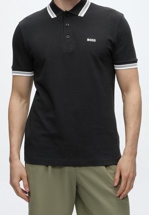 Mężczyzna noszący czarną koszulkę polo z białym wykończeniem na kołnierzu i rękawach oraz logo "BOSS" na lewej piersi, w połączeniu z oliwkowozielonymi spodniami.