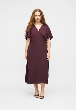 Kvinde stående vendt fremad iført en blommefarvet omslagskjole med flagrende ærmer og sorte åbne hæle på hvid baggrund.