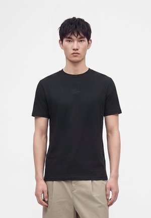 Ung mand med sort kort hår iført en ensfarvet sort T-shirt og beige bukser står mod en hvid baggrund.