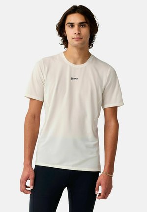 Giovane uomo con capelli scuri di media lunghezza che indossa una maglietta bianca con il logo Brooks e pantaloni scuri, in piedi davanti a uno sfondo semplice.