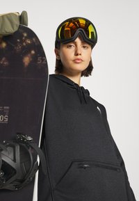 Čierna mikina s kapucňou a zipsom, ktorú nosí osoba držiaca snowboard. Snowboard má tmavú úpravu a čierne viazania.