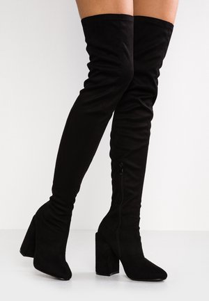 Bottes noires au-dessus du genou à talons blocs avec fermeture éclair latérale, portées aux jambes d'une personne sur un fond clair uni.
