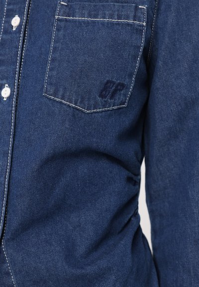 Chemise en denim bleu foncé avec une finition texturée. Présente une poche avant, des boutons blancs et des coutures blanches contrastantes. Logo sur la poche.