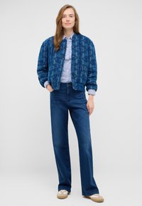 Niebieska bomberka w wzory, jasnoniebieska koszula w paski oraz ciemnoniebieskie szerokie spodnie dżinsowe. Brązowe buty typu slip-on z białymi akcentami.