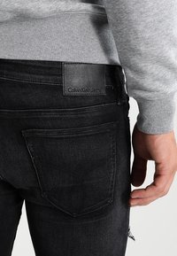 Vaqueros de mezclilla negra con una etiqueta de cuero que dice "Calvin Klein Jeans". Costuras visibles y bolsillos traseros con un patrón sutil. Sudadera gris encima.