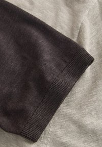 Nahaufnahme eines schwarzen Strickärmels mit geripptem Bündchen, der auf einem hellgrauen Strickstoff liegt.