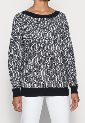 Kobieta w czarno-białym swetrze z geometrycznym wzorem z prążkowanymi ściągaczami na rękawach i u dołu, połączonym z białymi spodniami.