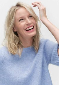 Ljusblå, mjuk stickad tröja med rund halsringning och korta ärmar. Modell med långt, vågigt blont hår som ler mot en neutral bakgrund.