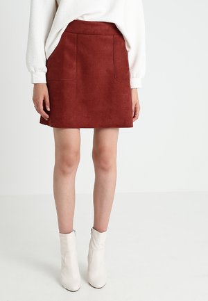 Vrouw draagt een roestkleurige suède minirok met voorzakken, een witte, gestructureerde top met lange mouwen en witte enkellaarzen, staand op een witte vloer.