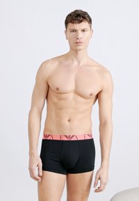 Άνδρας χωρίς πουκάμισο με κοντά σγουρά μαλλιά, που φοράει μαύρα μπόξερ με ροζ ζωνάκι με λογότυπο, στέκεται μπροστά σε απλό φωτεινό φόντο.