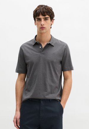 Jonge man die tegen een witte achtergrond staat, gekleed in een grijs poloshirt met korte mouwen en donkere broek, met één hand in de zak.