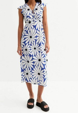 Kobieta w niebieskiej sukience midi z dużymi białymi stokrotkami z czarnymi środkami, w połączeniu z czarnymi sandałami na platformie z odkrytymi palcami.