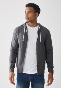 Young man wearing a gray zip-up hoodie over a white shirt and blue jeans, looking to the side against a plain light background.