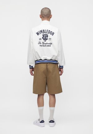 Personne à la tête rasée portant une veste blanche Wimbledon, un short marron, des chaussettes blanches et des baskets blanches Polo Ralph Lauren, debout, dos à nous.