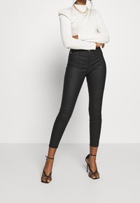 Jeans negros de tiro alto con un corte ajustado, hechos de mezclilla. Combinados con una blusa ceñida color crema y tacones negros con tiras. Detalle de collar de cadena dorada.