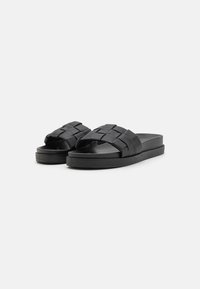 Black slide sandals featuring woven strap design, flat sole, and smooth texture. Minimalist style with a contemporary look.
