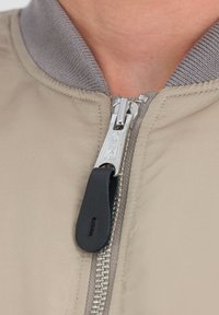 Chaqueta bomber beige con un collar acanalado gris, que presenta un zipper plateado y una pestaña de zipper negra. Textura suave y diseño minimalista.
