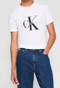 Biała, krótka koszulka z krótkim rękawem z dużym czarnym napisem "cK" oraz logo Calvin Klein Jeans, zestawiona z niebieskimi dżinsami, z jedną ręką w kieszeni.