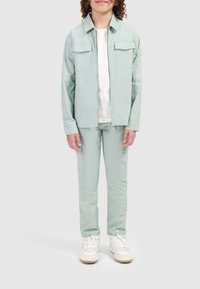 Lichtgroene jas met twee zakken aan de voorkant, gecombineerd met bijpassende broeken. Daaronder een wit shirt. Schoeisel bestaat uit witte sneakers met beige zolen.