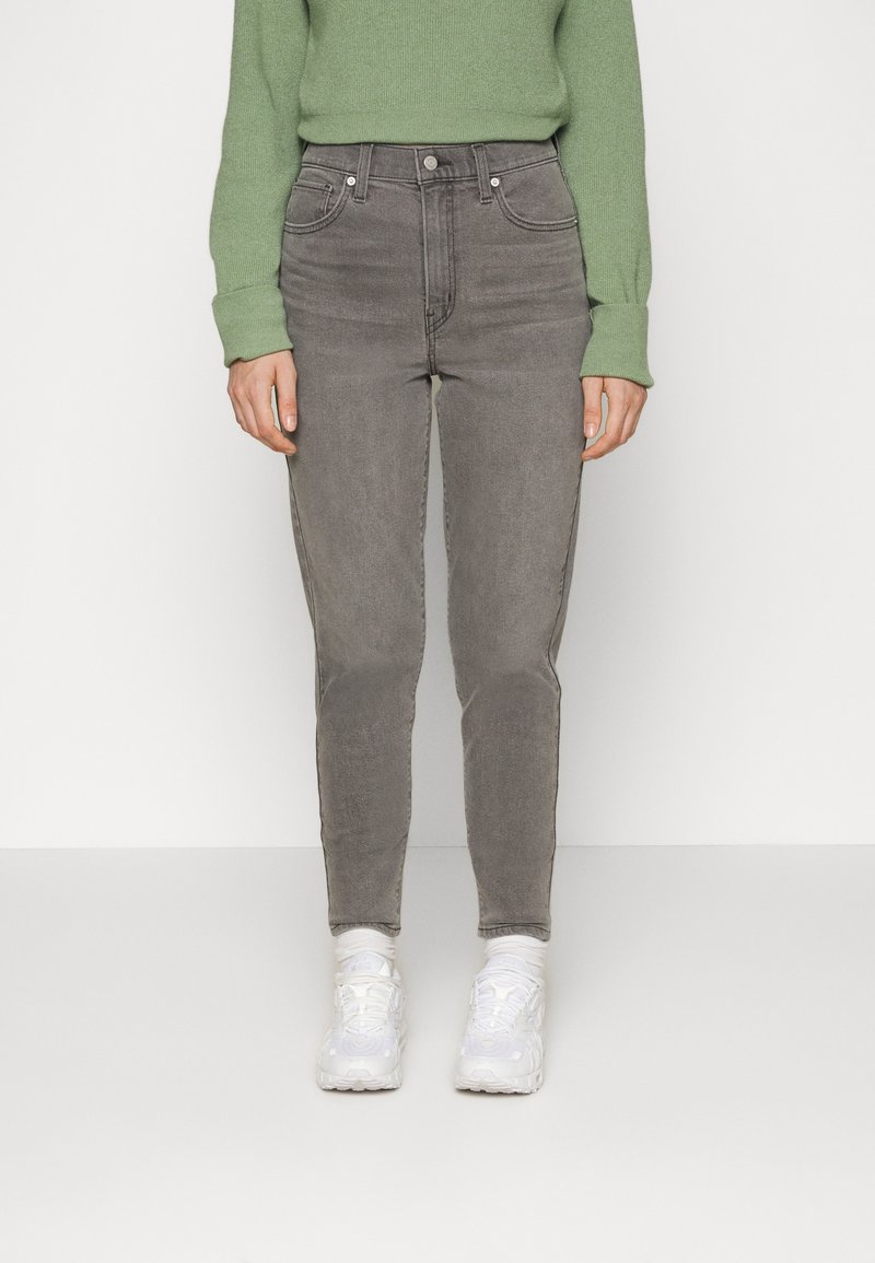 Persona con pantalones vaqueros de cintura alta grises, suéter verde, calcetines blancos y zapatillas blancas de pie contra un fondo liso.