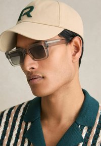 Beige cap with green "R," clear square sunglasses, and a color-blocked knit shirt featuring brown and teal patterns.