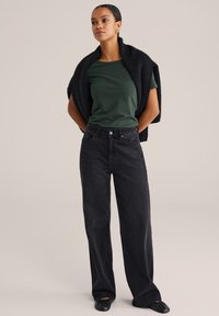 Melnas platās bikses saskarsē ar tumši zaļu īso piedurkņu blūzi, virsū uzlikta melna adīta šalle. Gluda tekstūra un ikdienišķs piegriezums.