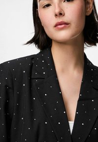 Juodas švarkas su struktūrizuotu apykaklė, dekoruotas išsklaidytais metaliniais smeigėmis, pasižymintis jų lygiu paviršiumi ir pritaikytu siluetu.