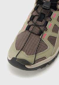 Közeli kép barna hálós túracipőről bézs borításokkal, fekete gyorsfűző rendszerrel és kis rózsaszín varrási részletekkel az oldalsó paneleken.