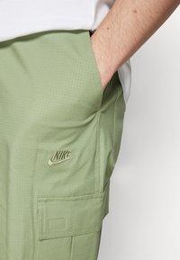 Ljusgröna cargobyxor med ett texturerat, rutnätsmönstrat mönster. Funktioner inkluderar sidofickor och en broderad Nike-logotyp i en mörkare grön färg.