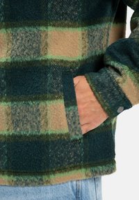 Flanellen jas met groen, bruin en donkerblauw ruitpatroon, met een zachte textuur, voorzak en drukknoopdetail op de manchetten.