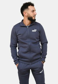Tuta con zip di colore blu navy dal design aderente. Presenta un piccolo logo bianco Puma sul petto e sui pantaloni. Tessuto morbido e testurizzato.