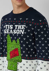 Granatowy sweter dzianinowy z białym napisem "'TIS THE SEASON", zielonym obrazkiem pudełka na prezent oraz czerwonymi akcentami i świątecznymi wzorami na całej powierzchni.