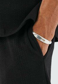 Una pulsera de bangles plateada descansa sobre una mano, con una superficie lisa y el nombre de la marca grabado. El fondo muestra una tela negra texturizada.