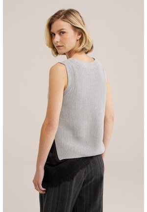 Donna con capelli biondi corti che indossa un top in maglia grigio chiaro senza maniche e pantaloni scuri a righe sottili con dettagli in pizzo, che guarda oltre la spalla.