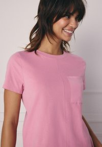 Roze t-shirt met korte mouwen van zachte katoen. Heeft een ronde hals en een klein borstzakje met een eenvoudig ontwerp en een gladde textuur.