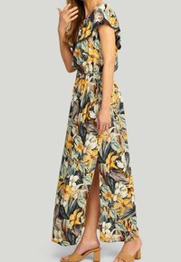 Maxikjole i blomsterprint med gule og grønne mønstre, off-the-shoulder flæser og en sideslids. Glat stof, ankel-længde.